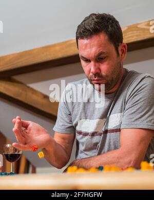 Young male adult is playing a board game.  Dice are seen in the air, showing two sixes mid air while falling down on a table. Glass of wine in the bac Stock Photo