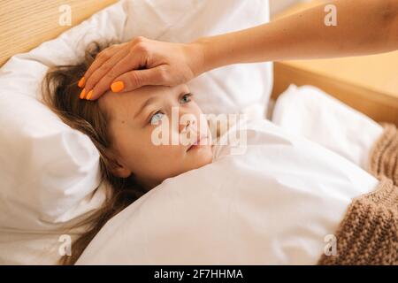 Close-up view of young mother measures temperature touches forehead of sick daughter lying in bed at home. 