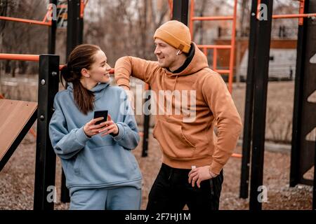 Young attractive sport woman and man in sportswear are smiling. Female is holding phone and male with AirPods, standing at sports park Stock Photo