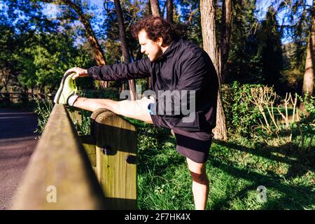 young adult man stretching the hamstrings on a wooden railing in a wooded park at sunset point concept personal care stretching and doing sports Stock Photo