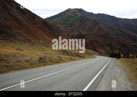 The asphalt road turns around the foot of the mountain, running through a beautiful valley in the mountains in autumn. Chuisky tract, Altai, Siberia, Stock Photo
