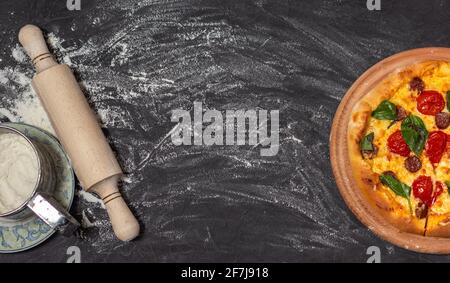 Pizza on wooden plate on black stone background. Rolling-pin and floor. Cheese pizza with tomato, mozzarella and basil. Banner. Close-up photo. Copy Stock Photo