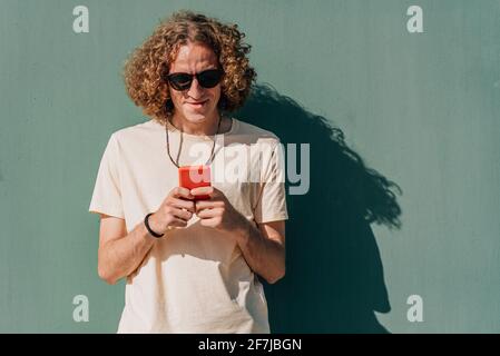 horizontal portrait of a young caucasian man using a red smart phone. He is dressing casual and rests near a green wall Stock Photo
