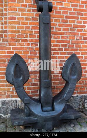 A huge antique black metal anchor with two large pointed legs standing on a gray stone and cobbled scaffold, propped up and attached to a red brick wa Stock Photo