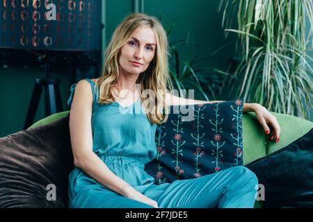 Beautiful graceful white woman is sitting on the sofa. Green living room interior. Relaxation. Stylish blonde. Stock Photo