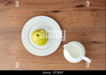 Top view of green pear on a white ceramic plate and empty milk jug on wooden table Stock Photo