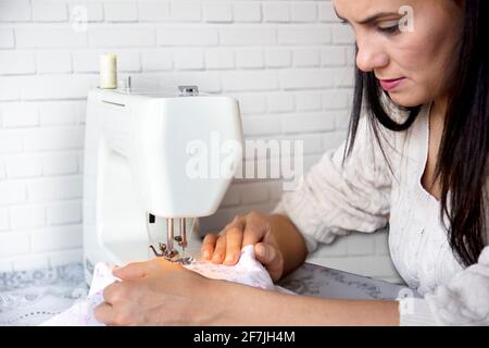 beautiful brunette young woman sews dress with the sewing machine. sewing at home as a hobby. selective Focus machine and hands. Stock Photo