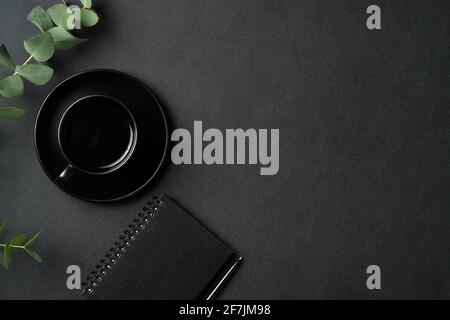 Coffee break concept. Black office desk table with cup of coffee, paper notebook, pen, eucalyptus on leather background. Flat lay, top view, copy spac Stock Photo