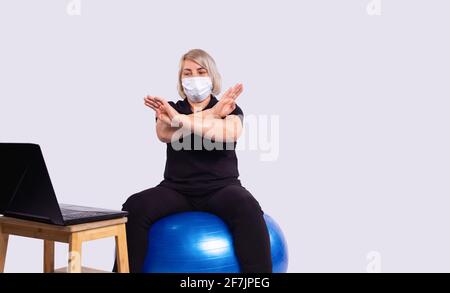 Senior blonde woman, fitness coach in medical mask on quarantine gesturing stop sign with hands looking at laptop, sport exercising, working from Stock Photo