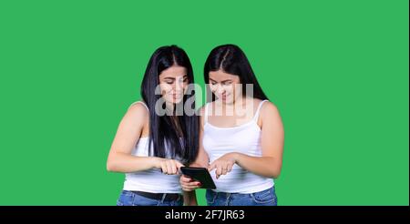 Banner, long format on green.Young smiling brunette twin sisters playing on digital tablet at home with copy space. Caucasian american women, cheerful Stock Photo