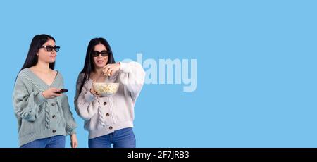 Banner, long format on blue background. Cute sisters, twins, girls wearing 3d glasses watching movie in evening, eating popcorn. Cinema show. Side Stock Photo