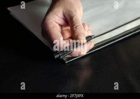 The officer's hands are holding a pen, a file folder on a black desk in close-up. A middle-aged man in a green camouflage uniform. Indoors. Selective Stock Photo