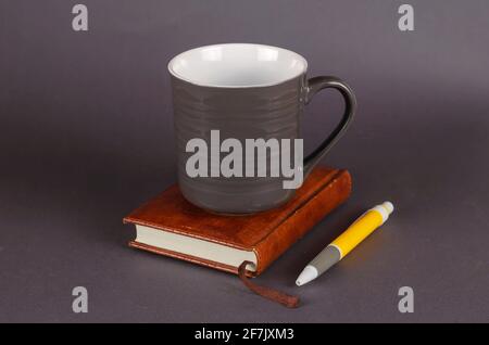 Empty ceramic mug, pen and notebook on gray background. New dark gray mug, yellow pen and brown notebook. Selective focus. Stock Photo