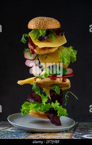 Levitating burger slices. Flying food. Hamburger bun, double chicken cutlet with cheese, lettuce, arugula and radish. Meat, vegetables and bread. Ingredients on black background. Stock Photo