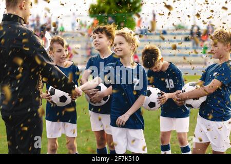 Happy Kids Celebrating Sports Success. Young Coach Congratulating to Boy Winning Football Soccer Tournament and Handing Out Reward - Golden Trophy. Sp Stock Photo