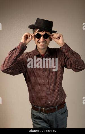 Brutal macho man in hat and round sunglasses standing isolated on studio copy space background. Trendy fashion young caucasian guy smiling touching ey Stock Photo