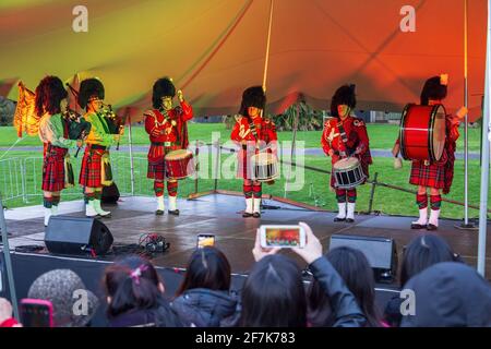 A Scottish pipe and drum band in red uniforms, kilts and busby hats, performing in a park in Auckland, New Zealand Stock Photo