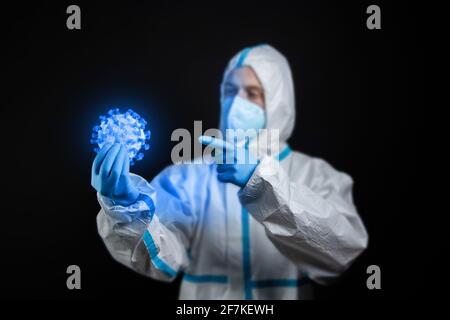 A young male doctor in a protective suit, a respirator and medical gloves holds a blue glowing corona virus during the coronavirus or Covid-19 pandemi Stock Photo