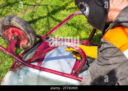 A man in a protective helmet welds a metal cart with an electrode. Agricultural machinery repair. Stock Photo