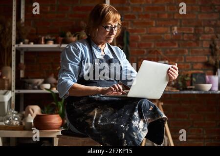Nice old woman using modern laptop in pottery workshop Stock Photo