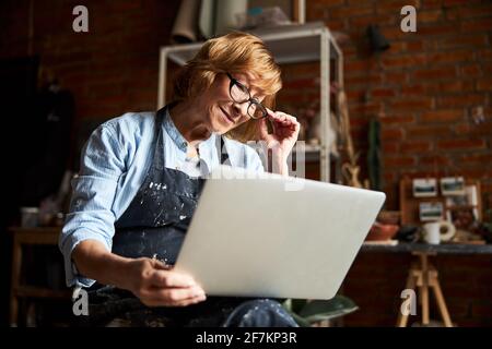 Joyful senior woman using laptop in pottery workshop Stock Photo