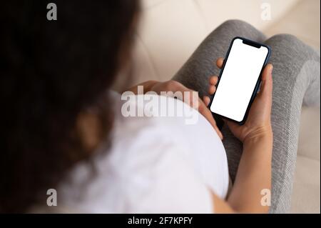 Wireless gadget, technology concept. Over shoulder view of pregnant woman in white t-shirt holding smartphone with blank white screen in hand, place for your presentation Stock Photo