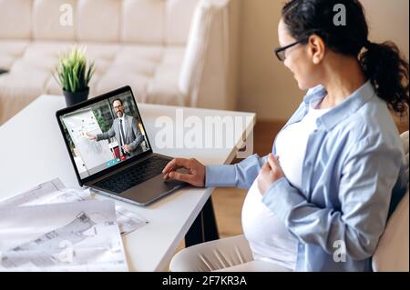 Virtual business meeting. Pregnant woman sitting at a desk, communicates on a video call using a laptop, remote work or distance learning, a male business colleague or tutor on the laptop screen Stock Photo