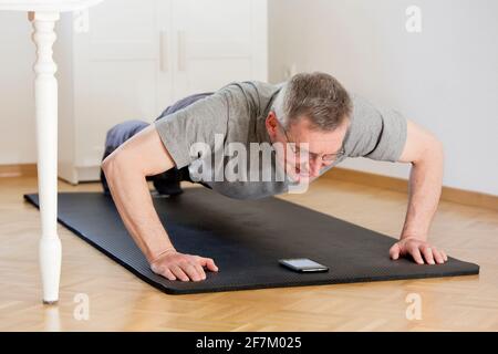 Mature man doing push ups at home with smart phone on the mat - selective focus on the face Stock Photo
