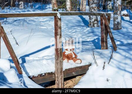 A stuffed toy teddy bear wearing a scarf and a hat sits on snow in the winter forest on a sunny day. Stock Photo