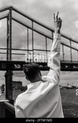 The guy with the raised hand shows the pis gesture on the background of the bridge. Black and white photo. Stock Photo
