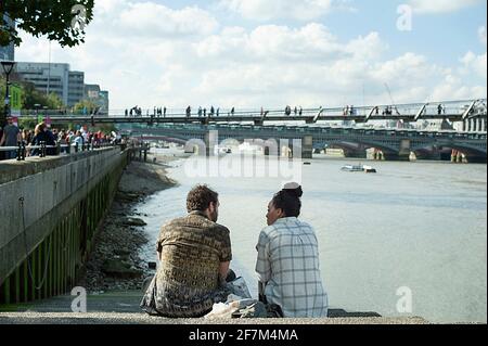 Rear view of 30s young friends or couple talking by the stairs, Bankside. Iconic location with the Millennium bridge, Thames river, London, UK Stock Photo