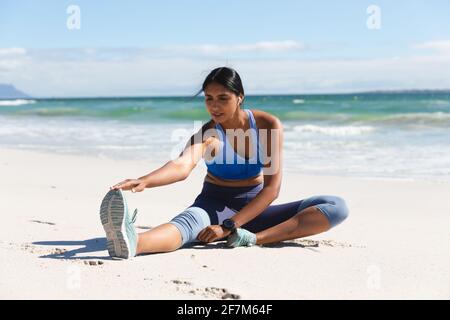 Mixed race woman exercising on beach wearing wireless earphones sitting and stretching Stock Photo