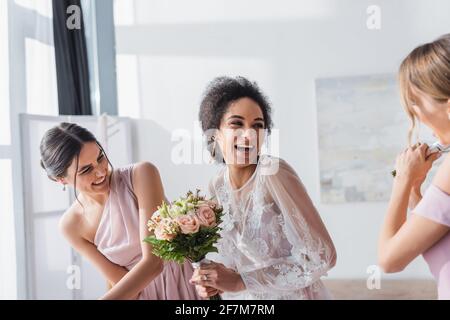 excited african american bride holding wedding bouquet and laughing near bridesmaids Stock Photo