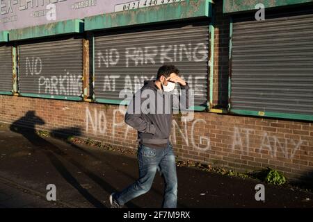 Man wearing a face mask walking past no parking signs painted on a wall and shutters to prevent pavement parking in Balsall Heath on 7th January 2021 in Birmingham, United Kingdom. Birmingham has become a city where locals say that a certain irresponsibility has developed, where drivers show little respect for the law, and seem to park anywhere they choose. Stock Photo