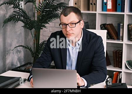 Serious man in glasses and a business suit looks at the camera while sitting at an office table and a laptop on the background of a rack with folders Stock Photo