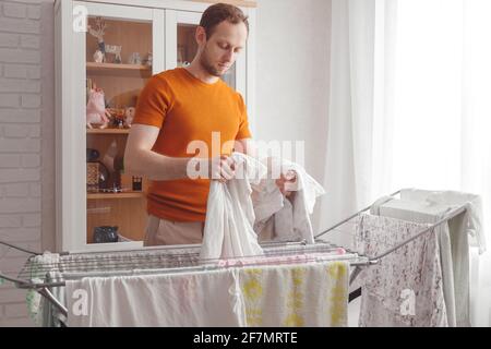 Man doing home chores. Caucasian man removes clothing and baby sheets after laundry from portable dryer in living room Stock Photo