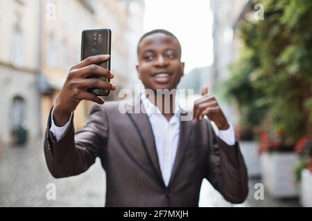 Happy afro american businessman using modern smartphone for video chat outdoors. Handsome man in stylish suit having online conversation on street. Stock Photo