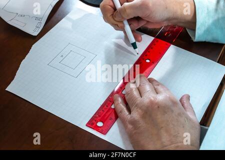 A woman uses a pen to count measurements and sketch out a floor plan for a house on square graph paper using a clear red ruler on hardwood desk. Stock Photo