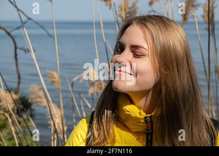 Portret of happy smiling young woman with her eyes closed enjoys the sunshine and fresh air. She stands in against lake on warm spring day in yellow Stock Photo