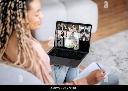Distance learning, online lecture. Over shoulder view of african american female student sitting on sofa with laptop, taking notes and listening online lesson, teacher and students on laptop screen Stock Photo