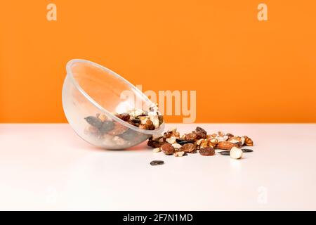 Various nuts spilled from a cup on the white table against bright orange background. Selective focus, minimalistic, vivid colors. Stock Photo