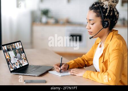 Online webinar, business meeting. Side view of african american woman, communicates with business colleagues by video call uses laptop, takes notes, multiracial coworkers on laptop screen, brainstorm Stock Photo