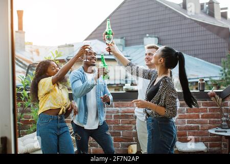 Multi ethnic group of friends dancing and relaxing on rooftop. Group young hipsters drinking alcohol and chatting. Carefree weekends. Stock Photo