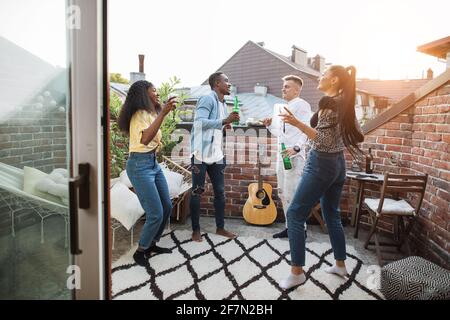 Multicultural people with alcoholic drinks in hands dancing and singing. Cheerful young friends in casual wear enjoying party time on open terrace.