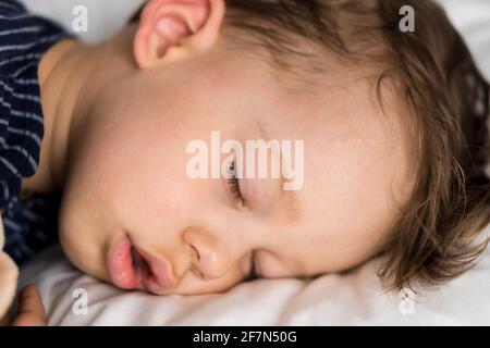 Authentic portrait sick cute caucasian little preschool baby boy in blue sleep with teddy bear on white bed. child resting at lunchtime. care Stock Photo