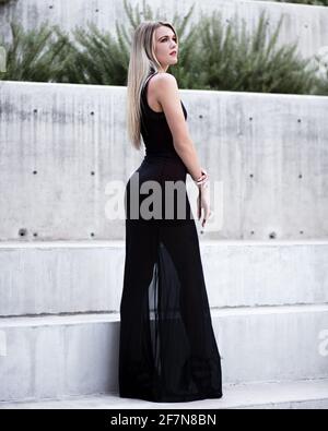 Blonde girl wearing a long black dress posing in a urban scene with cement walls in the background. Fashionable and lifestyle concept. Stock Photo