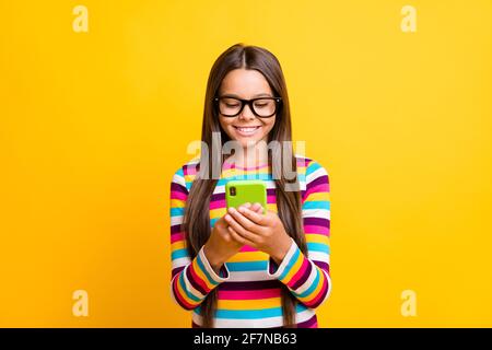 Photo of small girl hold telephone look screen white smile wear specs striped shirt isolated yellow color background Stock Photo