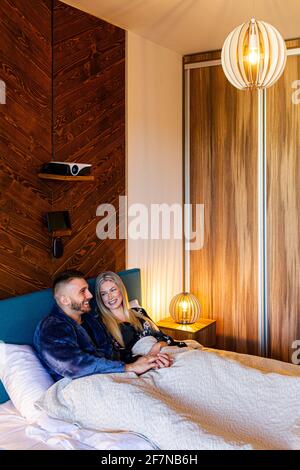 A young smiling couple semi-sitting (semi-lying) in bed in bedroom, covered to the waist with a blanket. Stock Photo