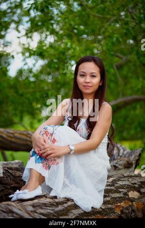 Full body adult filipino female in dress looking at camera while relaxing on trunk of tree on summer day in forest Stock Photo