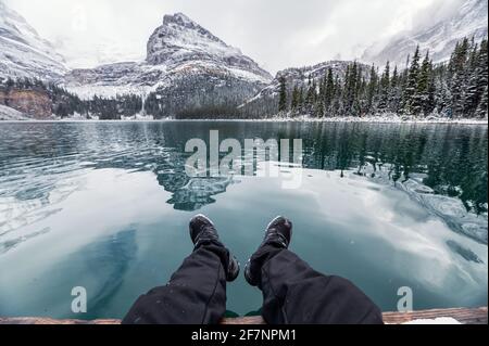 Legs of traveler relaxing on pier with rocky mountains reflection in Lake O'hara on winter at Yoho National Park, Canada Stock Photo